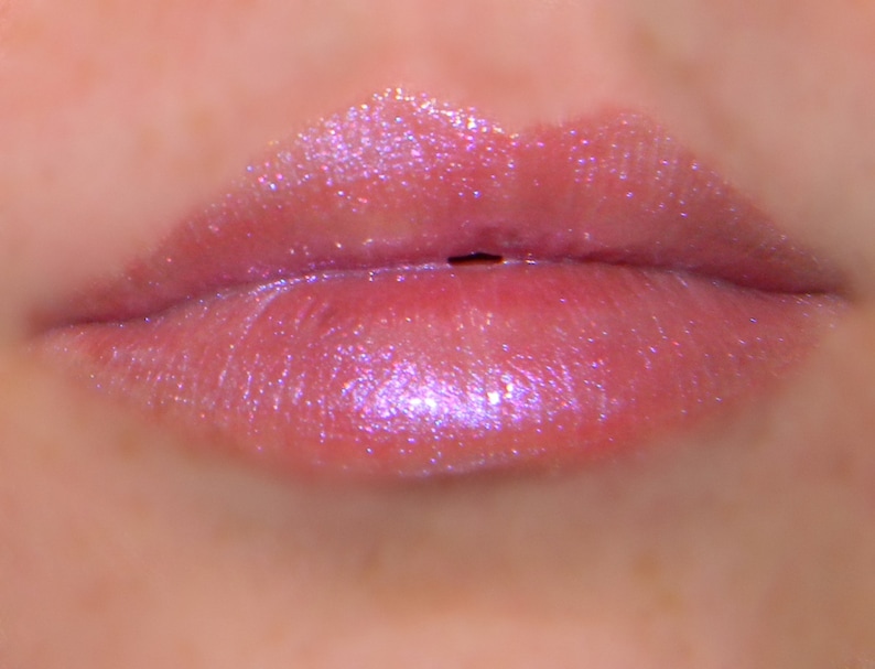 May include: Close-up of pink lips with a clear, iridescent lip gloss. The gloss has a subtle shimmer and reflects light.