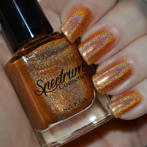 May include: A bottle of Spectrum Cosmetics nail polish in a gold holographic color. The bottle is clear and shows the polish inside. The polish is a fine shimmery gold with a rainbow effect.