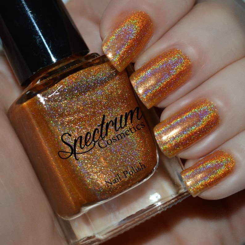 May include: A bottle of Spectrum Cosmetics nail polish in a gold holographic color. The bottle is clear and shows the polish inside. The polish is a fine shimmery gold with a rainbow effect.