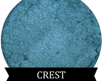 May include: A teal blue eyeshadow with a shimmery finish. The eyeshadow is in a round container with the word "CREST" printed on a black ribbon.