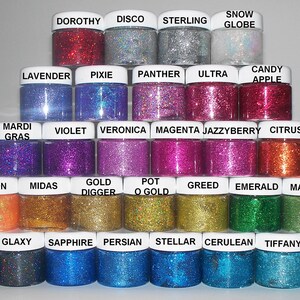 May include: A collection of 24 small jars of glitter, arranged in a pyramid shape. Each jar has a different color of glitter and a label with a name. The names include Dorothy, Disco, Sterling, Snow Globe, Lavender, Pixie, Panther, Ultra, Candy Apple, Mardi Gras, Violet, Veronica, Magenta, Jazzyberry, Citrus, Melon, Midas, Gold Digger, Pot O Gold, Greed, Emerald, Mantis, Midnight, Glaxy, Sapphire, Persian, Stellar, Cerulean, Tiffany, and Mermaid.