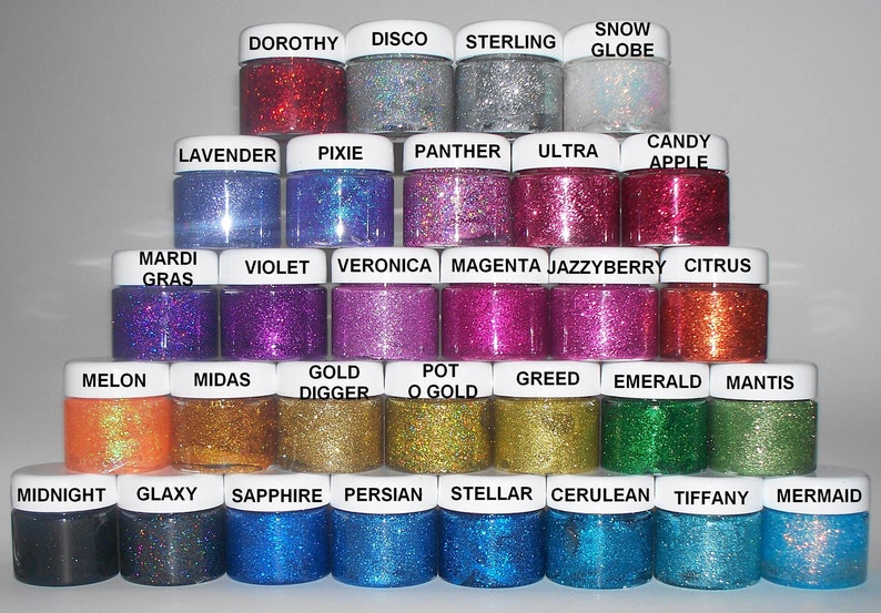 May include: A collection of 24 small jars of glitter, arranged in a pyramid shape. Each jar has a different color of glitter and a label with a name. The names include Dorothy, Disco, Sterling, Snow Globe, Lavender, Pixie, Panther, Ultra, Candy Apple, Mardi Gras, Violet, Veronica, Magenta, Jazzyberry, Citrus, Melon, Midas, Gold Digger, Pot O Gold, Greed, Emerald, Mantis, Midnight, Glaxy, Sapphire, Persian, Stellar, Cerulean, Tiffany, and Mermaid.