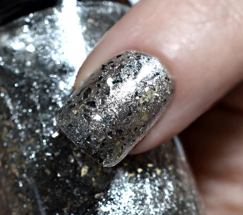 May include: Close-up of a fingernail painted with silver glitter nail polish. The polish has a mix of silver, black, and gold glitter flakes. The nail is held over a bottle of the same nail polish.