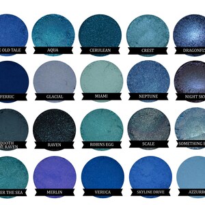 May include: A collection of 15 blue and green eyeshadows in round pans. The eyeshadows are arranged in a grid of five rows and three columns. The names of the eyeshadows are printed below each pan.
