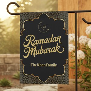 May include: A black garden flag with gold text that reads "Ramadan Mubarak" and "The Khan Family." The flag has a gold decorative border and a crescent moon and star. The flag is displayed on a black metal garden stake.
