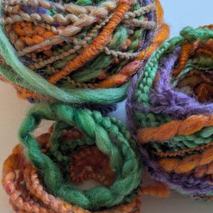May include: Three balls of art yarn featuring a blend of textures and colors including orange, lime green, forest green, and purple. The yarn consists of various plies, including thick, fuzzy, braided, and twisted strands wound together in spheres.