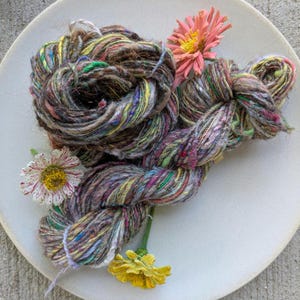 May include: A skein of multi-colored, textured yarn featuring a blend of brown, green, purple, and yellow fibers. The yarn is arranged in a loose coil and is decorated with three fresh flowers: a pink zinnia, a white and red striped zinnia, and a yellow zinnia.