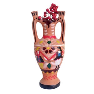 Op de afbeelding: Een terracotta vaas met twee handvatten en een geschilderd ontwerp. De vaas heeft een rood, wit en geel patroon en illustraties van dansende mensen. De vaas is gevuld met rode bessen.