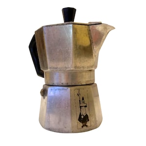 Puede incluir: Una cafetera italiana plateada con asa y tapa negras. La cafetera tiene un diseño octogonal clásico y una pequeña pegatina con una figura de dibujos animados. La cafetera está diseñada para preparar espresso.