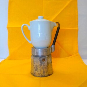 May include: A white ceramic coffee pot with a lid sits atop a silver and gold stovetop espresso maker. The pot has a handle and spout. The espresso maker has a black handle. The background is a bright yellow cloth.
