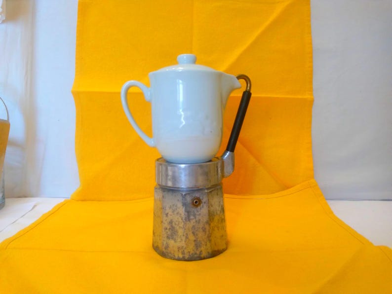 May include: A white ceramic coffee pot with a lid sits atop a silver and gold stovetop espresso maker. The pot has a handle and spout. The espresso maker has a black handle. The background is a bright yellow cloth.