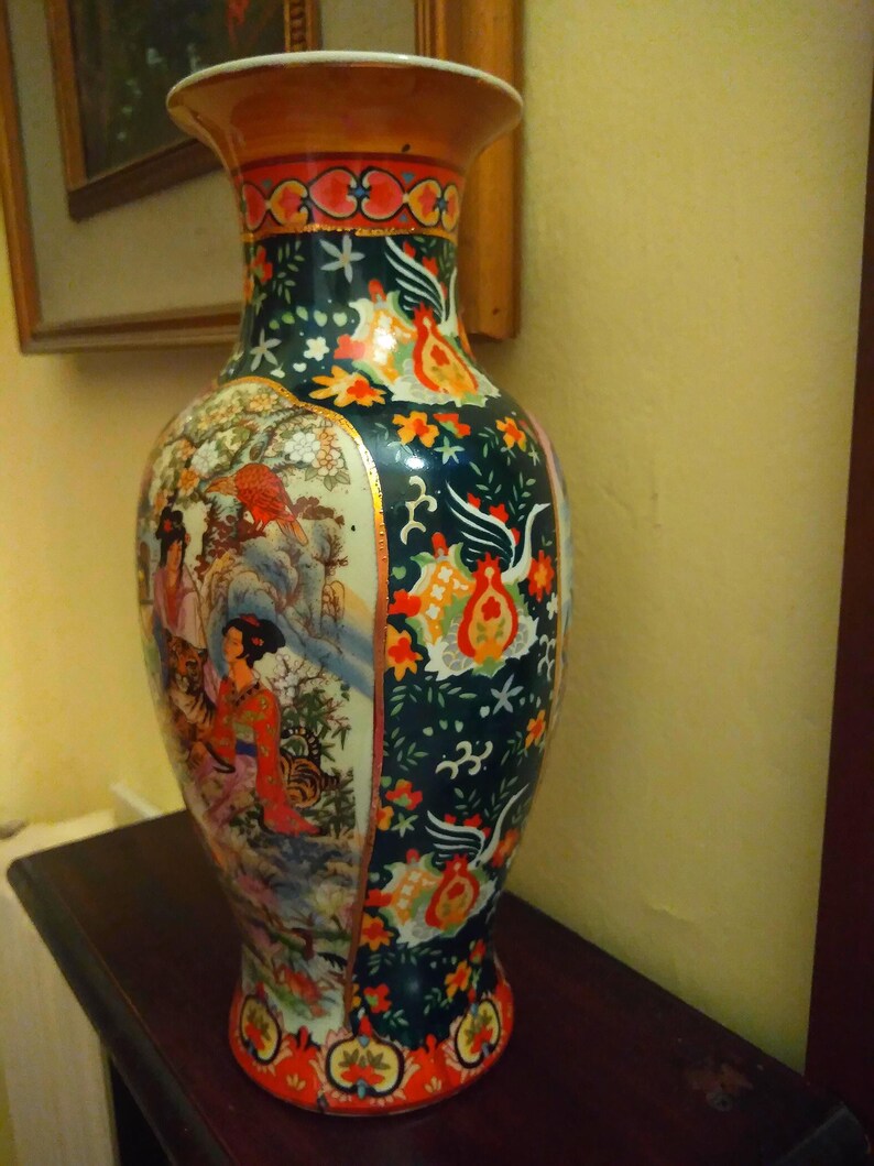 Pu&ograve; includere: Un vaso di ceramica alto e ornato con una base ampia e un bordo svasato. Il vaso presenta uno sfondo verde scuro con motivi floreali e di uccelli, e un pannello raffigurante figure in un paesaggio. Il bordo &egrave; marrone con dettagli rossi e dorati.