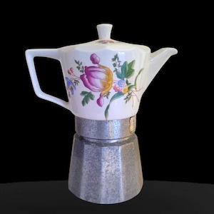 Peut inclure: Une cafetière italienne blanche en céramique avec un motif floral comprenant des fleurs violettes, jaunes et bleues. La base est en métal argenté. La poignée et le bec sont blancs.