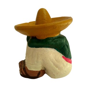 May include: A small, painted figurine of a person wearing a yellow sombrero and a green jacket. The figure is in a seated position, with white pants and brown shoes. A red sash is visible.
