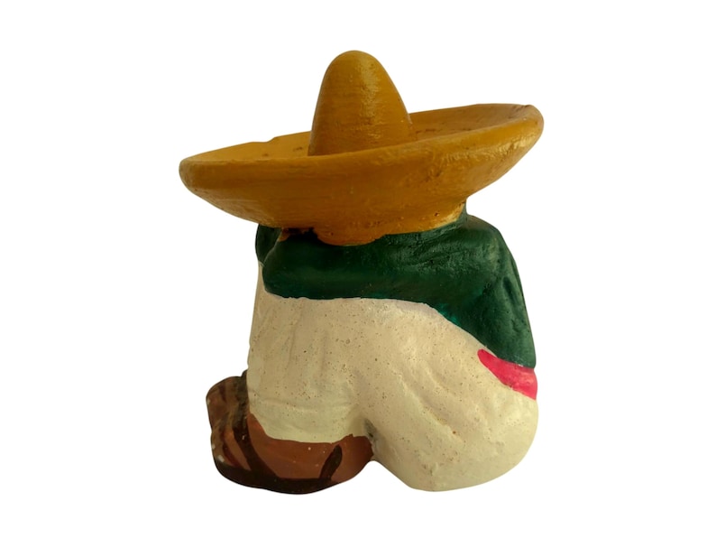 May include: A small, painted figurine of a person wearing a yellow sombrero and a green jacket. The figure is in a seated position, with white pants and brown shoes. A red sash is visible.