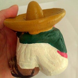 May include: A hand-painted figurine of a person wearing a sombrero. The figure is dressed in a green jacket, white pants, and a red sash. The sombrero is tan, and the figure's shoes are brown.