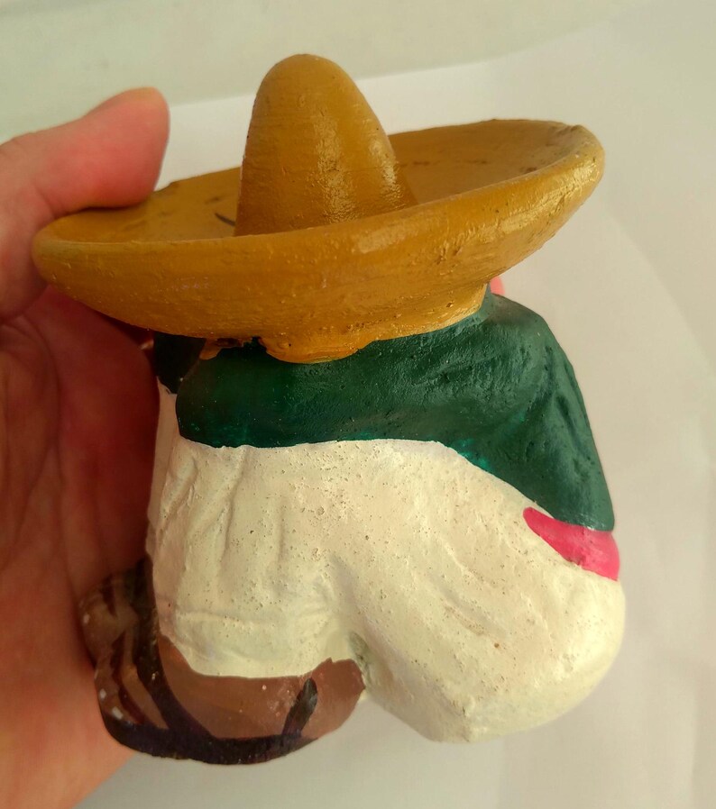 May include: A hand-painted figurine of a person wearing a sombrero. The figure is dressed in a green jacket, white pants, and a red sash. The sombrero is tan, and the figure's shoes are brown.