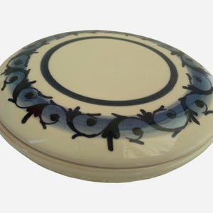 Könnte beinhalten: Ovale Schmuckschatulle aus Keramik mit cremefarbenem Boden und Deckel. Der Deckel hat einen dunkelblauen Rand und ein dekoratives blaues Blumenmuster am Rand. Die Mitte des Deckels ist heller cremefarben.