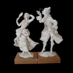 Könnte beinhalten: Zwei cremeweiße Keramikfiguren, die ein Paar in historischer Kleidung darstellen, jeweils auf einem quadratischen Holzsockel montiert. Eine Figur hält einen Handspiegel, die andere eine Brille. Die Skulpturen zeigen detaillierte Kleiderstrukturen.