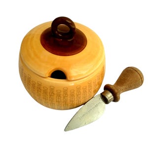Könnte beinhalten: Ein hellgelber Käsebehälter mit einem dunkelbraunen Deckel und einem Käsemesser. Der Behälter ist mit "Parmigiano Reggiano" beschriftet. Das Messer hat einen Holzgriff und eine silberne Klinge.
