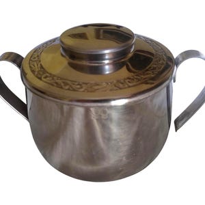May include: A stainless steel sugar bowl with a lid and two curved handles. The lid has a decorative embossed design around the edge. The bowl is round and shiny, reflecting the light.