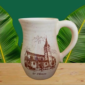 Op de afbeelding: Crèmekleurige keramische kan met een bruine illustratie van een kerk en de tekst "St. Chéron". De kan heeft een grote handgreep en een brede opening. Groene bladeren op de achtergrond.
