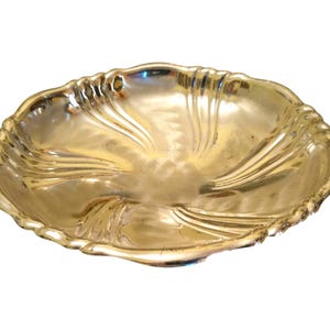 Puede incluir: Un plato decorativo de color dorado con un borde festoneado. El plato tiene un diseño arremolinado y texturizado que irradia desde el centro. El plato es probablemente de metal y está sobre un fondo blanco.