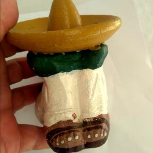 May include: A small, hand-painted figurine of a person wearing a yellow sombrero and a green and white outfit. The figurine has brown shoes with white dots. The figure is held by a hand.