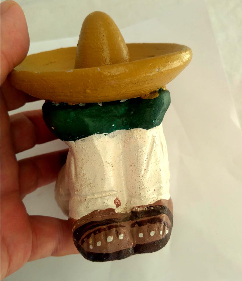 May include: A small, hand-painted figurine of a person wearing a yellow sombrero and a green and white outfit. The figurine has brown shoes with white dots. The figure is held by a hand.