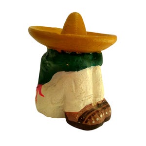 May include: A ceramic figurine of a person wearing a yellow sombrero and a green and white outfit. The figurine has brown shoes and a red accent. The figure is painted with a textured finish.