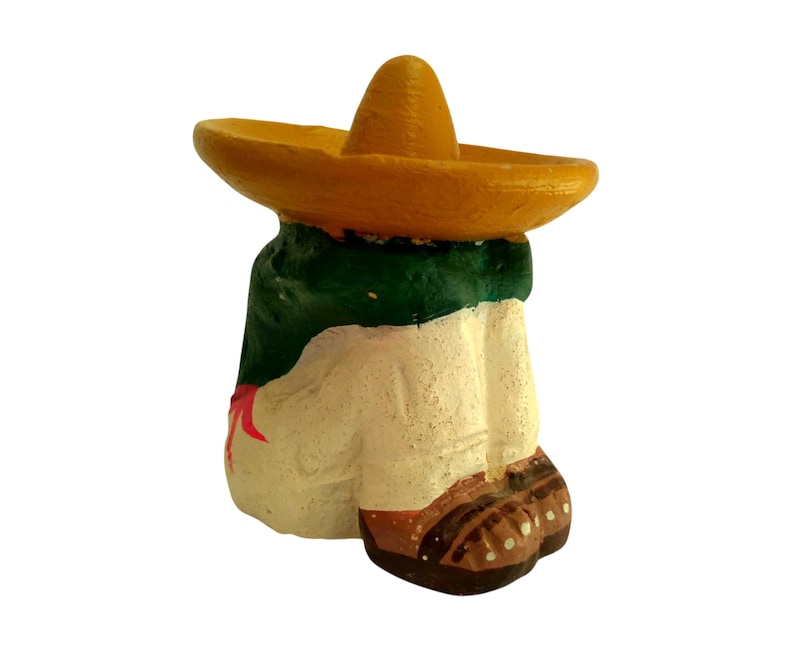May include: A ceramic figurine of a person wearing a yellow sombrero and a green and white outfit. The figurine has brown shoes and a red accent. The figure is painted with a textured finish.