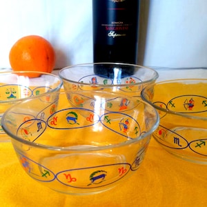 May include: Four clear glass bowls decorated with zodiac signs in blue, yellow, and red. They sit on a yellow surface, with an orange and a dark bottle in the background. These bowls are likely for serving food.