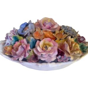 Può includere: Una composizione floreale in ceramica in un piatto bianco a forma di conchiglia. I fiori sono un mix di colori pastello, tra cui rosa, giallo, blu e arancione. Il piatto ha un bordo strutturato e smerlato.