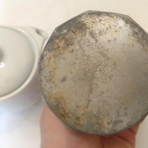 May include: Close-up of a white ceramic lid and a silver-colored, octagonal metal object. The metal object has a worn, aged appearance with visible rust and discoloration. The lid is part of a small, white ceramic dish.