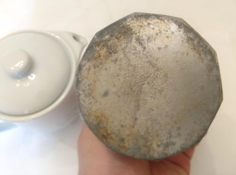 May include: Close-up of a white ceramic lid and a silver-colored, octagonal metal object. The metal object has a worn, aged appearance with visible rust and discoloration. The lid is part of a small, white ceramic dish.