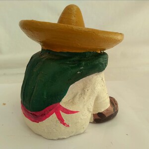 May include: A ceramic figurine of a person wearing a yellow sombrero and a green poncho. The figure is painted with white pants, a red sash, and brown shoes. The figurine is in a seated position.