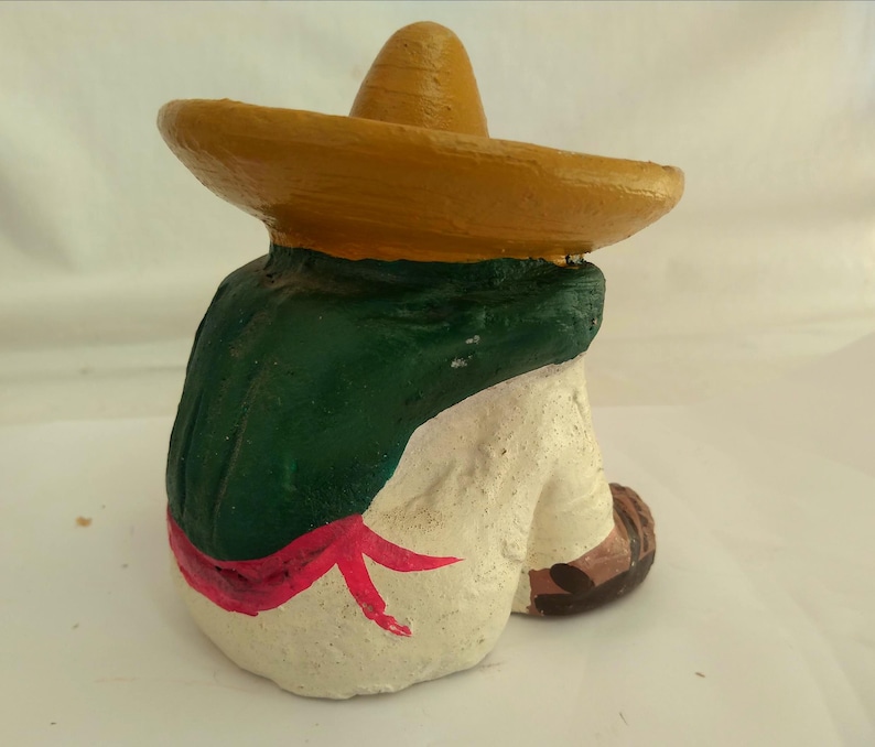 May include: A ceramic figurine of a person wearing a yellow sombrero and a green poncho. The figure is painted with white pants, a red sash, and brown shoes. The figurine is in a seated position.