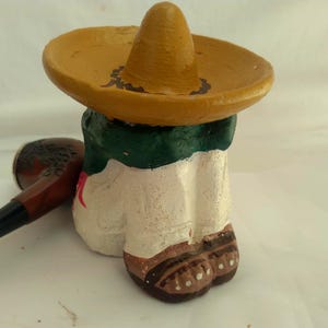 May include: A hand-painted ceramic figurine of a person wearing a yellow sombrero and a green and white outfit. A brown pipe rests beside the figurine. The figurine's shoes are brown with white dots.