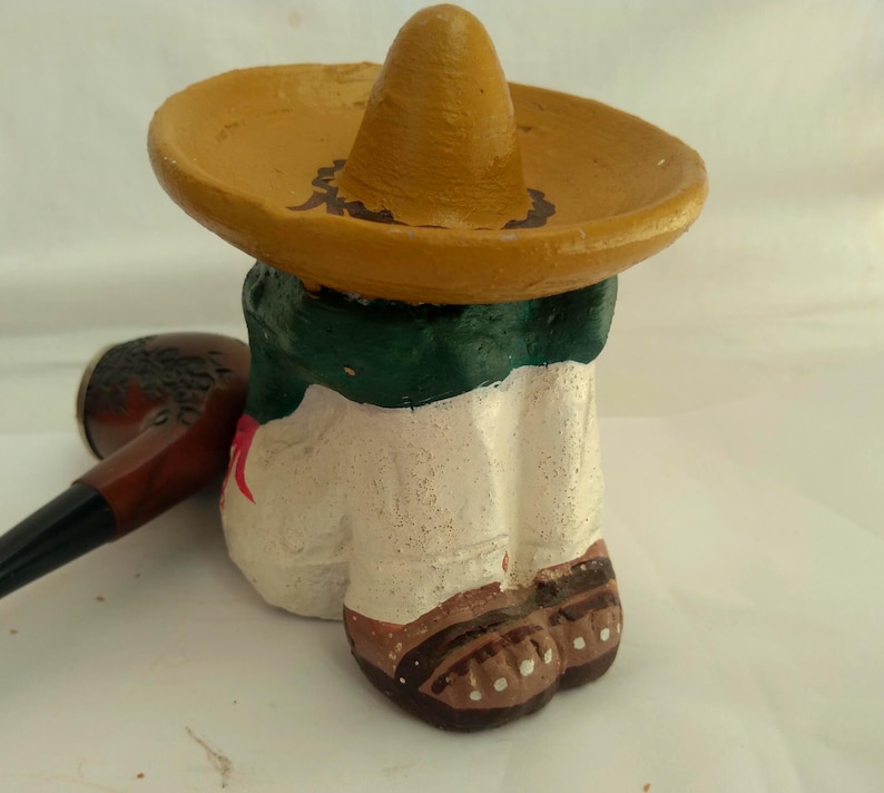 May include: A hand-painted ceramic figurine of a person wearing a yellow sombrero and a green and white outfit. A brown pipe rests beside the figurine. The figurine's shoes are brown with white dots.
