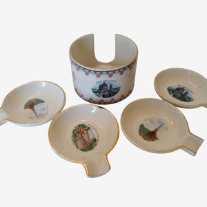 May include: A set of four small white ceramic ashtrays with gold-trimmed edges and a matching cylindrical holder. Each ashtray and the holder feature colorful illustrations of Italian landmarks, including the Mole Antonelliana in Turin.