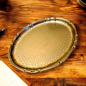 Puede incluir: Una bandeja ovalada plateada con una superficie martillada y bordes festoneados decorativos. La bandeja está hecha de un metal brillante y se coloca sobre un fondo de madera cálida. Una pieza clásica para servir alimentos o bebidas.