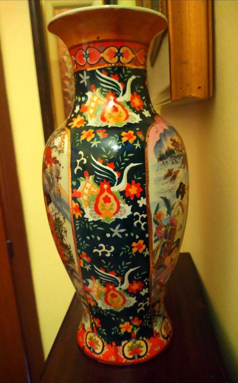 Pu&ograve; includere: Un vaso decorativo in ceramica alto con una base larga e un bordo svasato. Il vaso presenta uno sfondo verde scuro con uccelli bianchi, motivi floreali arancioni e rossi e scene di persone e paesaggi. Il bordo &egrave; marrone e rosso.