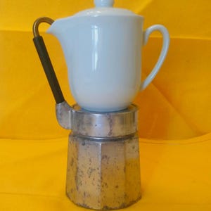 May include: A vintage stovetop espresso maker with a white ceramic top and a silver-colored base. The handle is black with a gold-colored accent. The espresso maker is on a yellow background.