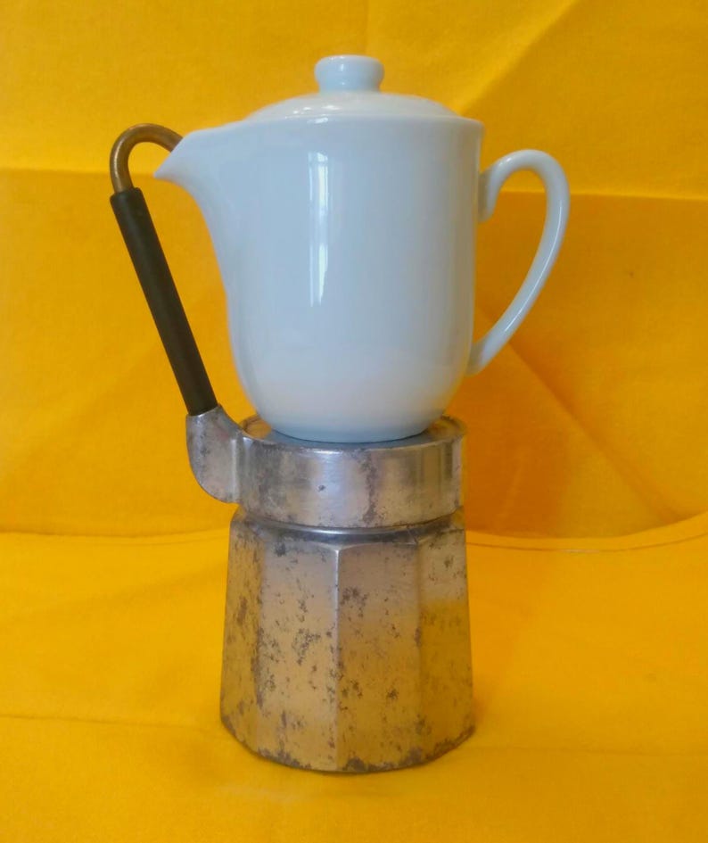 May include: A vintage stovetop espresso maker with a white ceramic top and a silver-colored base. The handle is black with a gold-colored accent. The espresso maker is on a yellow background.