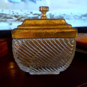 Puede incluir: Un recipiente cuadrado de vidrio con un patrón texturizado de costillas diagonales y una tapa metálica extraíble. La tapa presenta un acabado dorado desgastado y un pequeño pomo cilíndrico central para levantarla. La base de vidrio es transparente.