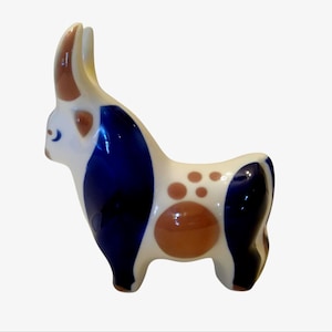Może przedstawiać: Ceramiczna figurka byka z białą podstawą, ozdobiona niebieskimi i brązowymi akcentami. Byk ma zakrzywiony róg i zaokrąglony korpus, z brązowymi plamami i dużym brązowym okręgiem z boku. Przedmiot dekoracyjny.