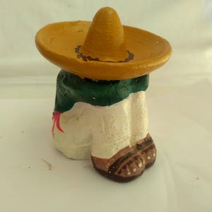 May include: A painted figurine of a person wearing a sombrero. The figure has a yellow hat, a green and white torso, and brown shoes. A red bow is visible on the front of the figure.