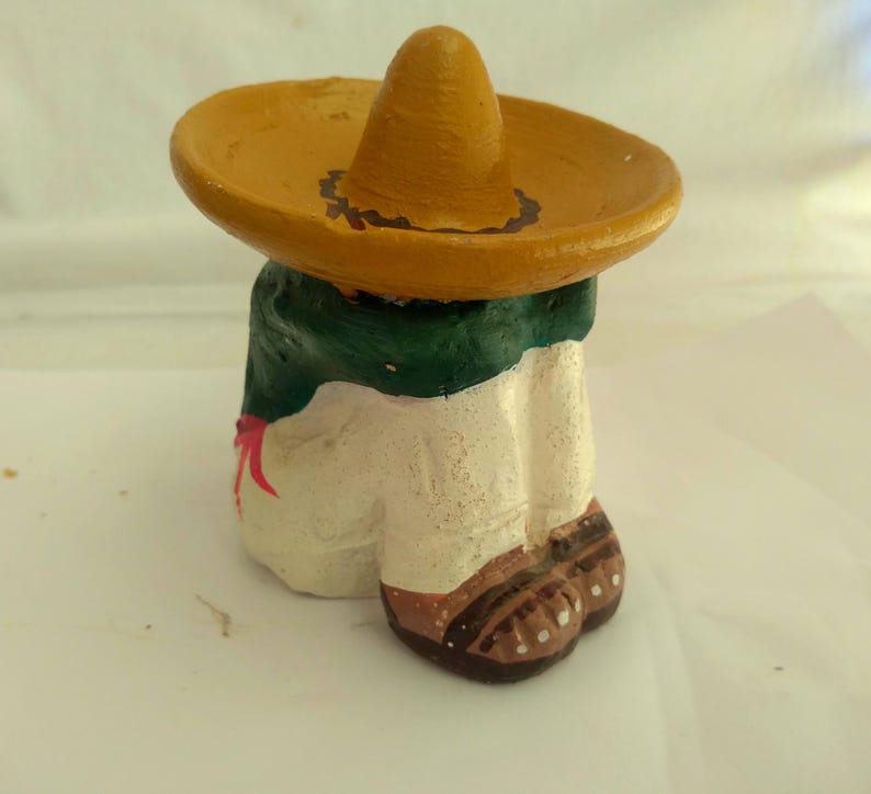 May include: A painted figurine of a person wearing a sombrero. The figure has a yellow hat, a green and white torso, and brown shoes. A red bow is visible on the front of the figure.