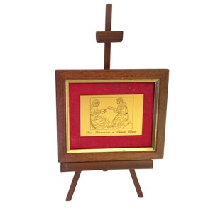 May include: A framed gold-colored religious image on a small wooden easel. The image features two figures with text below. The frame is brown with a red matting. The easel is also brown.