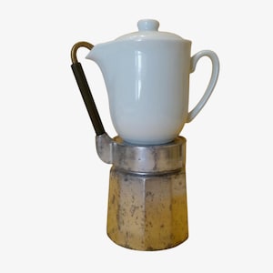 May include: A vintage stovetop espresso maker with a white ceramic top and a silver-colored base. The handle is black with a brass accent. The coffee maker is designed to brew espresso on a stovetop.